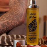 Kiwi Glow's Kavai Tattoo Oil sitting on a striped surface with a feather in the background. A mans torso and biceps are visible behind the product, though blurry.