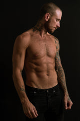 “Tattooed man with a bare chest standing in dim studio lighting, showcasing healed tattooed skin.”