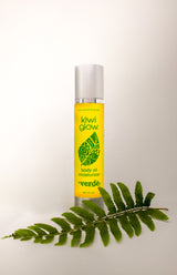 Kiwi Glow's Verde Body Oil sits in a white space with a green fern in the foreground. The bottle is yellow and has the Kiwi Glow Leaf Logo on the center area of the bottle. It reads: "Kiwi Glow... Body Oil Moisterizer... Verde...net 4 fl oz."