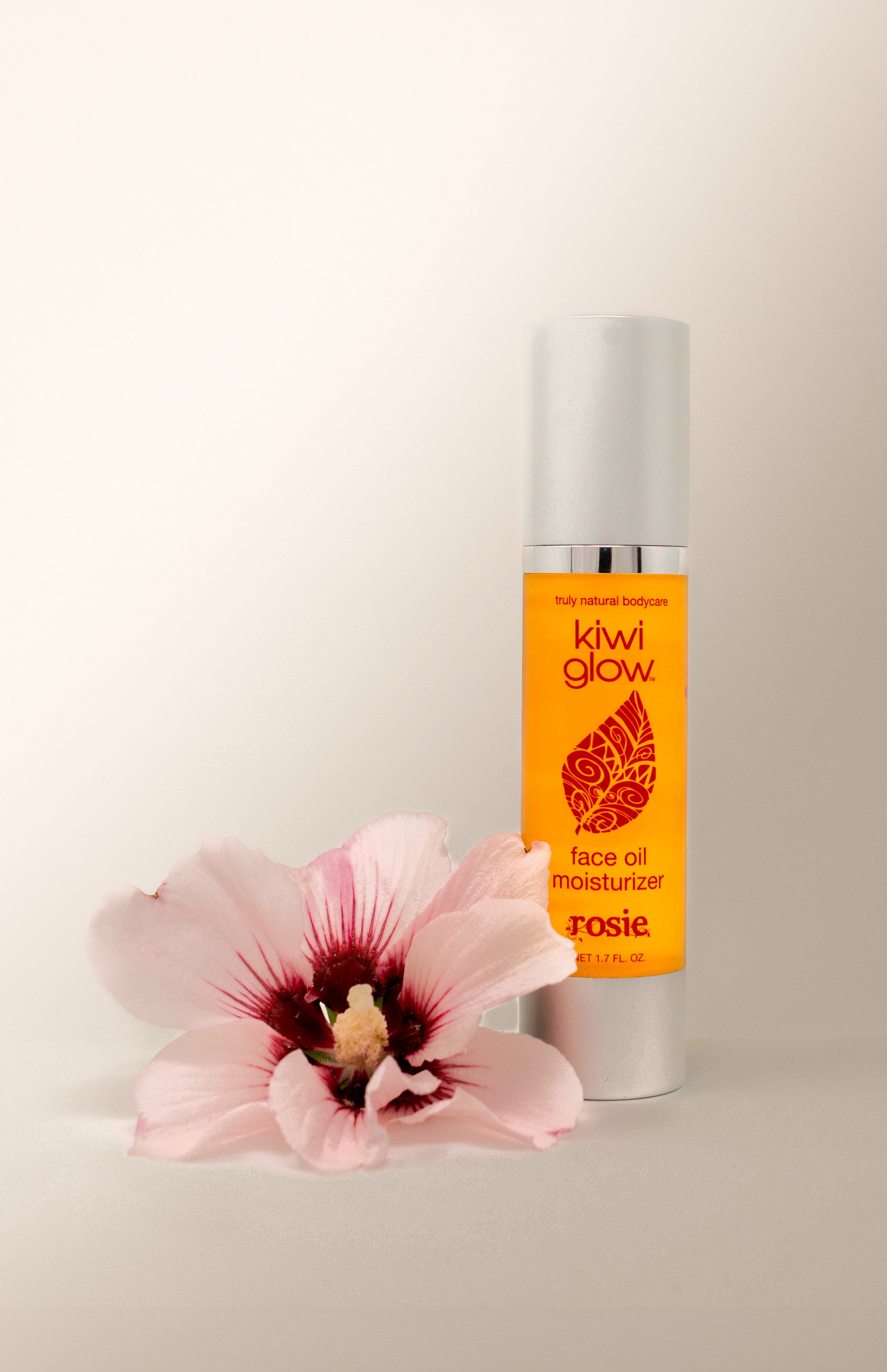 Kiwi Glow's Rosie Face Oil sits in a white space with a pink flower in the foreground. The bottle is yellow and has the Kiwi Glow Leaf Logo on the center area of the bottle. It reads: "Kiwi Glow... Face Oil Moisterizer... Rosie...net 1.7 fl oz."