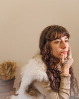 a young and beauitful woman rests her face in her right hand as if thinking about something. She is looking away from the camera and is wearing a white fur vest and a light brown sweater. She has long wavy deep brown hair and short. bangs. She is sitting against a white background.