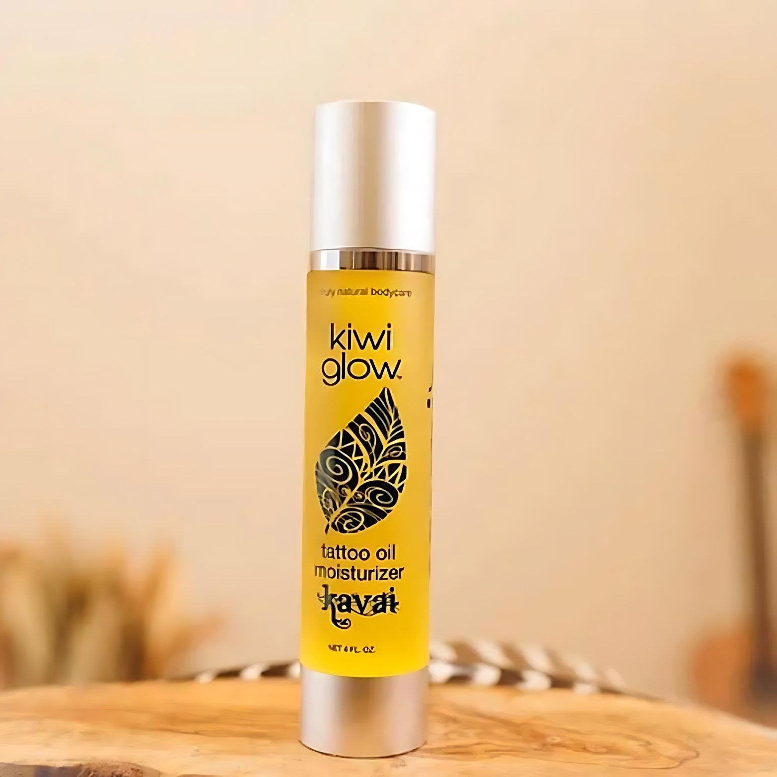 Kiwi Glow's Kavai Tattoo Oil sits on a wooden surface with a feather in the light brown background. The bottle is yellow and has the Kiwi Glow Leaf Logo on the center area of the bottle. It reads: "Kiwi Glow... Tattoo Oil Moisturizer... Kavai...net 4 fl oz."