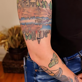 Man's bicep showing a tattoo of a castle and city with a river and squid. He is wearing a black shirt and blue jeans. Only his arm and torso are visible.