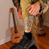 man's body in a blue and orange flannel and khaki cargo pants showing a shin and calf tattoo. foot is resting on a stool and a blurry guitar and wooden floor makes up the background. 